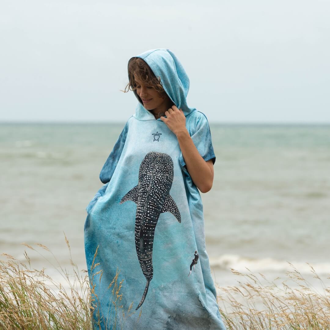 A woman wrapped in a light blue hooded towel poncho with a large whale shark design, standing on a beach with the ocean in the background.