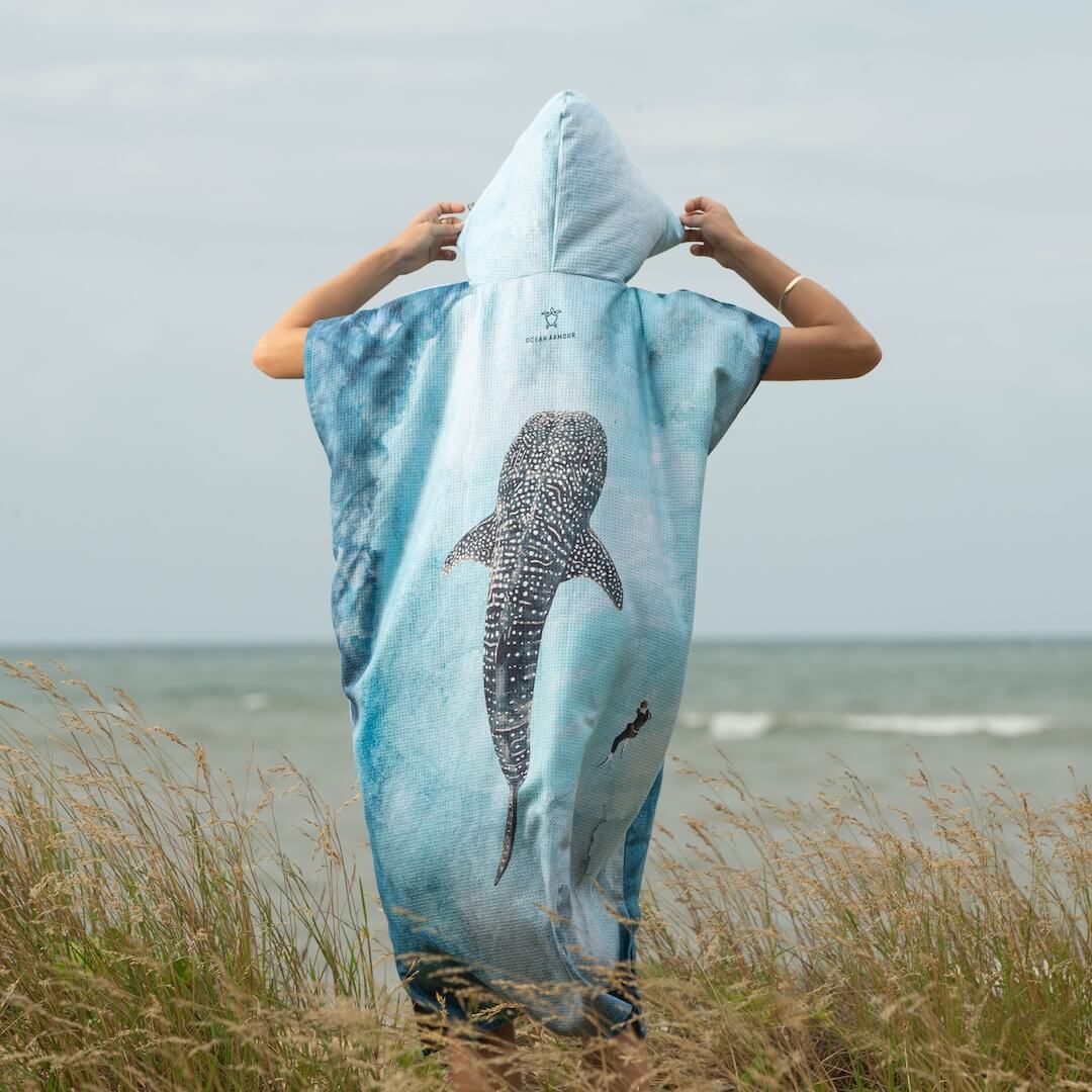 A person standing on grassy dunes facing the ocean, wearing a hooded towel poncho with a large watercolour whale shark print in shades of blue and white.